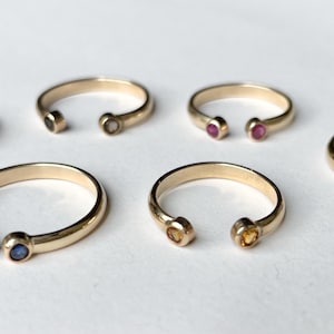 May include: Several gold-coloured rings, each with a small, colourful gemstone set at the open ends. The gemstones are in various colours, including green, clear, pink, blue, yellow, and purple. The rings are displayed on a white surface.