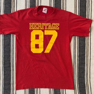 May include: A short-sleeved red t-shirt featuring the word HERITAGE in yellow block letters above the large yellow number 87. The shirt has a crew neckline and a white brand label visible on the inside of the collar at the back of the neck.