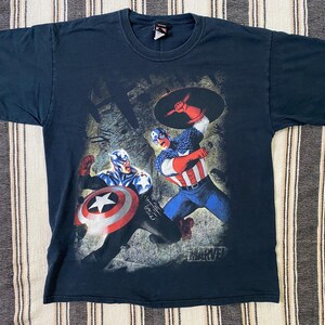 May include: Dark navy blue t-shirt featuring a graphic of Captain America in action, with his shield raised. The Marvel logo is visible at the bottom right. The shirt has a crew neck and short sleeves.