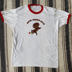 May include: A white ringer t-shirt with red trim on the collar and sleeves. The front features a brown graphic of a squirrel holding a small flower, with the text Alpha Gamma Delta arched in brown lettering above the squirrel illustration.