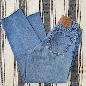 May include: Medium-wash blue denim jeans featuring a high-waisted fit, a classic five-pocket design, a tan leather brand patch on the back waistband, a small red tab on the back pocket, and raw, frayed edges at the bottom of the pant legs.