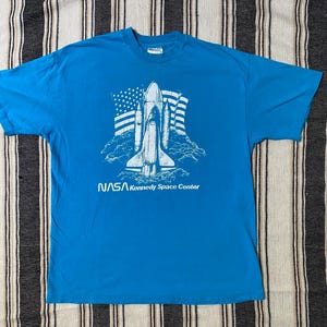 May include: A turquoise blue t-shirt featuring a white graphic of a space shuttle, the American flag, and the text "NASA Kennedy Space Center." The shirt is made of a soft material and has a crew neck.