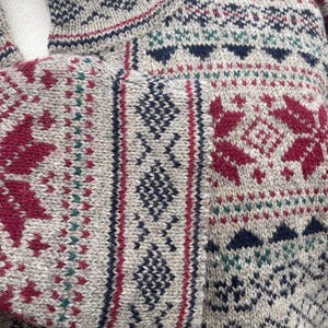 May include: Close-up of a beige knitted sweater with a high collar and patterned design. The sweater features burgundy, navy, and teal geometric and floral patterns. The sleeve cuff has a vertical stripe design.