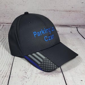 May include: Black baseball cap with the text "Parking Lot Czar" embroidered in blue. The brim features a design with gray stripes, a blue stripe, and a pattern of white stars.