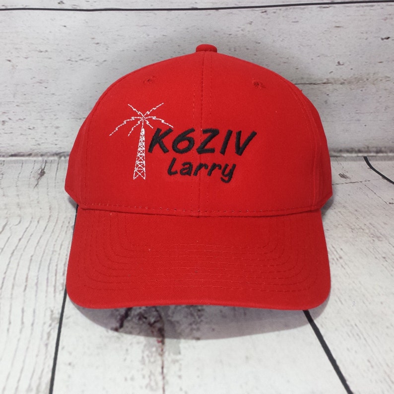 May include: A bright red baseball cap featuring an embroidered white palm tree graphic next to the black text K6ZIV and the name Larry embroidered in black script below the call sign on the front panel of the hat.