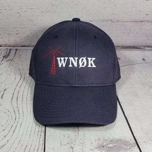 May include: A dark navy blue baseball cap featuring an embroidered design on the front. The embroidery includes a red palm tree graphic positioned to the left of the white text that reads WN&Oslash;K in a bold, capitalized, serif-style font.