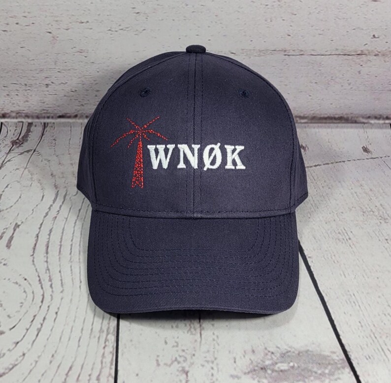 May include: A dark navy blue baseball cap featuring an embroidered design on the front. The embroidery includes a red palm tree graphic positioned to the left of the white text that reads WN&Oslash;K in a bold, capitalized, serif-style font.