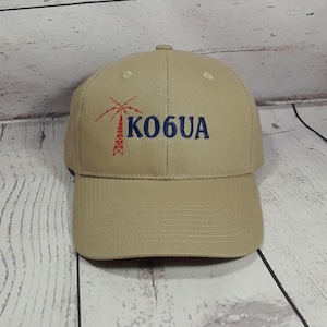 May include: A tan baseball cap featuring an embroidered red radio tower graphic and the blue text KO6UA on the front. The cap has a structured crown, a curved brim, and stitched eyelets on the top panels for ventilation and a classic fit.