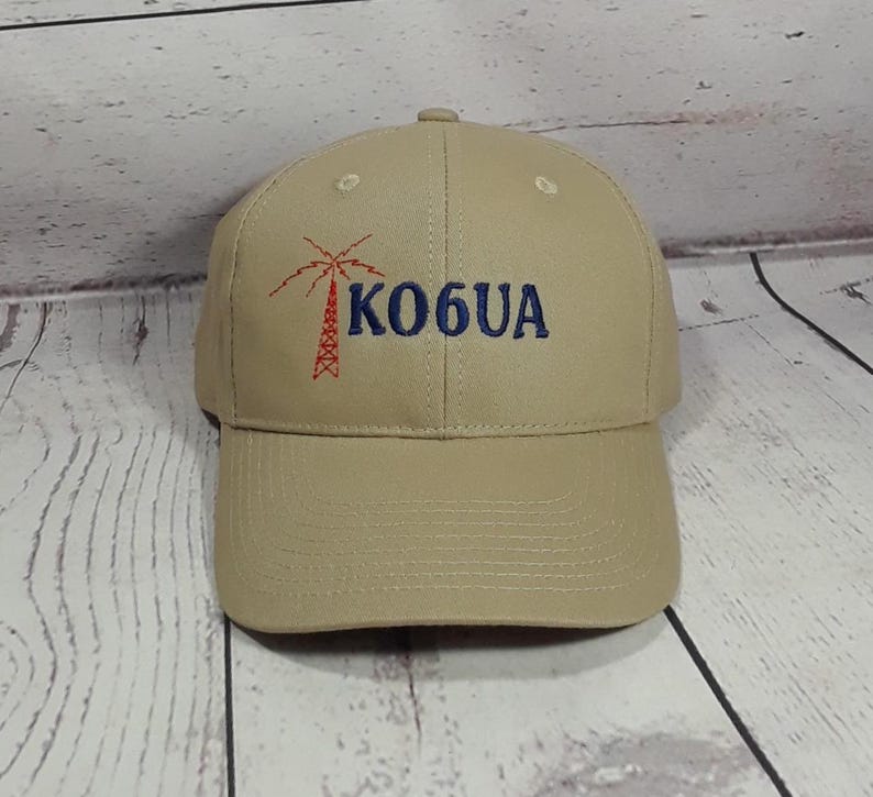 May include: A tan baseball cap featuring an embroidered red radio tower graphic and the blue text KO6UA on the front. The cap has a structured crown, a curved brim, and stitched eyelets on the top panels for ventilation and a classic fit.