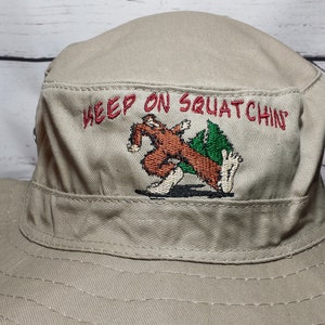 May include: A tan canvas bucket hat with an embroidered design of a brown Bigfoot-like creature walking with the text "Keep on Squatchin'" in red.
