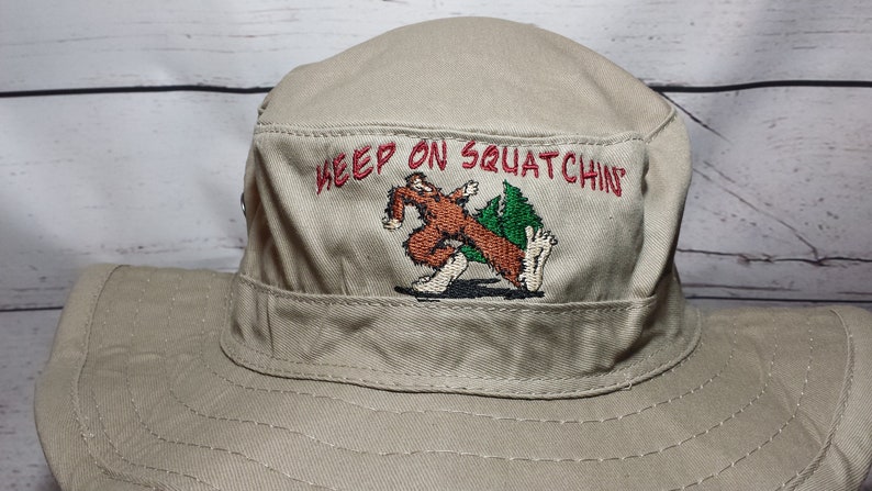 May include: A tan canvas bucket hat with an embroidered design of a brown Bigfoot-like creature walking with the text "Keep on Squatchin'" in red.