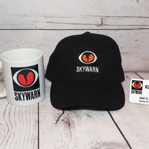 May include: A white ceramic mug, a black baseball cap, and a white name tag with the "SKYWARN" logo. The mug and cap feature a logo with a red heart inside an eye shape. The cap has "SKYWARN" embroidered on it.