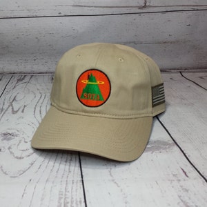 May include: A beige baseball cap with an embroidered patch featuring a green mountain with a red circle around it. The text "SOTA" is written in black on the patch. The cap has a small American flag patch on the side.
