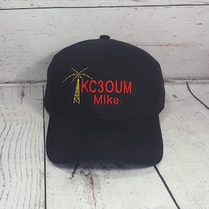 May include: A black baseball cap featuring an embroidered yellow radio tower icon on the left side, with the red text KC3OUM and the name Mike embroidered in red below the tower. The cap has a structured crown and a curved brim.