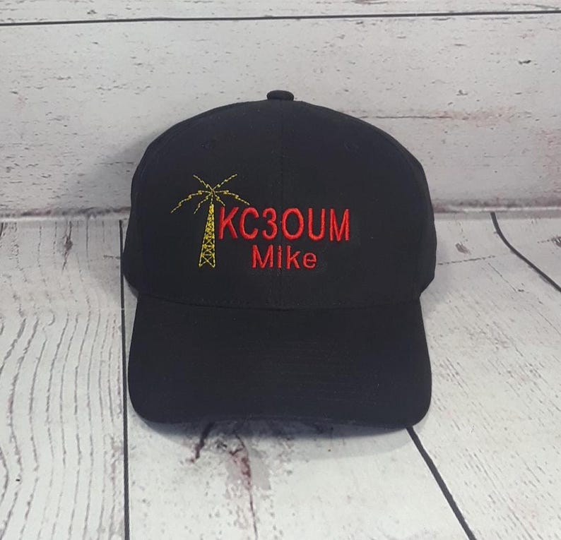 May include: A black baseball cap featuring an embroidered yellow radio tower icon on the left side, with the red text KC3OUM and the name Mike embroidered in red below the tower. The cap has a structured crown and a curved brim.