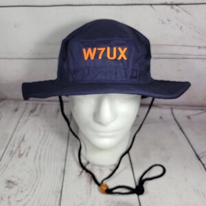 May include: Navy blue hat with a wide brim and the orange text "W7UX" on the front. The hat has a black chin strap with an adjustable toggle. The hat is displayed on a white mannequin head.
