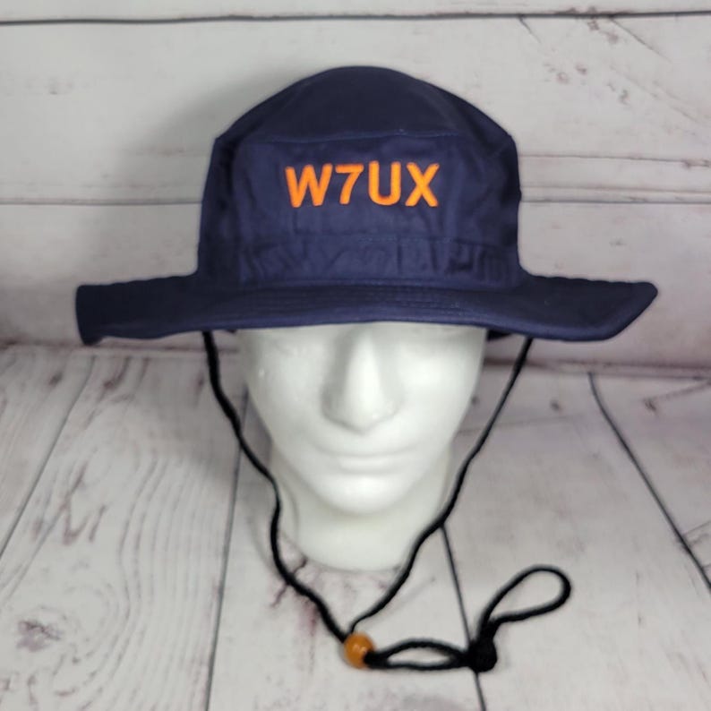May include: Navy blue hat with a wide brim and the orange text "W7UX" on the front. The hat has a black chin strap with an adjustable toggle. The hat is displayed on a white mannequin head.