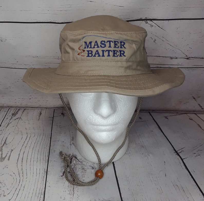 A khaki color, wide-brimmed sun hat with a khaki cord and a wooden bead. The hat has the words 'MASTER BAITER' embroidered on the front in navy blue thread along with a fish hook and worm
