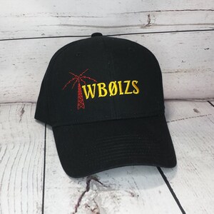 May include: A black baseball cap featuring an embroidered design on the front. The design includes a red radio tower icon next to the yellow embroidered text WB&Oslash;IZS. The cap has a structured crown, a curved brim, and a button on the top center.