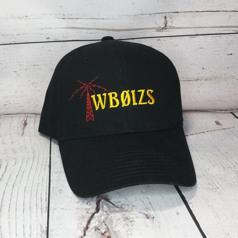 May include: A black baseball cap featuring an embroidered design on the front. The design includes a red radio tower icon next to the yellow embroidered text WB&Oslash;IZS. The cap has a structured crown, a curved brim, and a button on the top center.