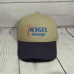 May include: A tan baseball cap with a dark navy blue brim. The front features an embroidered red radio tower icon next to the blue text W3GEL and the name George embroidered in blue below the call sign on the tan crown of the hat.