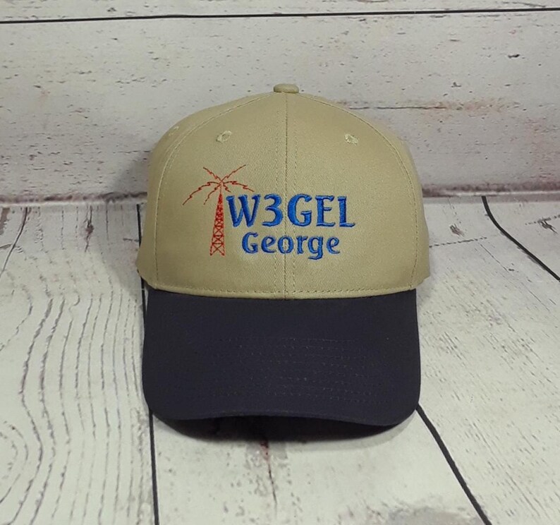May include: A tan baseball cap with a dark navy blue brim. The front features an embroidered red radio tower icon next to the blue text W3GEL and the name George embroidered in blue below the call sign on the tan crown of the hat.