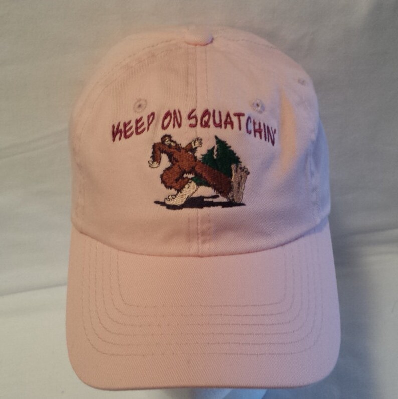 Op de afbeelding: Roze baseballpet met een geborduurd beeld van een Bigfoot die met een boom rent. De tekst "KEEP ON SQUATCHIN'" is in maroon geborduurd op de voorkant van de pet.