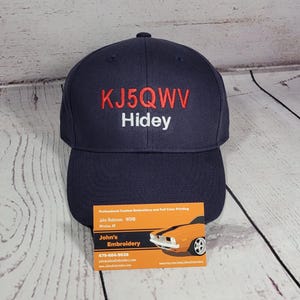 May include: A navy blue baseball cap featuring embroidered text on the front. The top line of text is in red and reads KJ5QWV, while the bottom line of text is in white and reads Hidey. The cap has a structured crown and a curved brim.