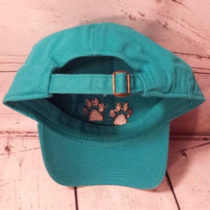 May include: Teal blue baseball cap with an adjustable strap and two white paw prints embroidered on the back.