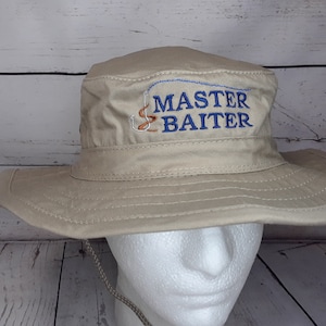 A khaki, wide-brimmed, bucket hat with a navy-blue text, a fish hook, and worm embroidered design that says 'MASTER BAITER'.