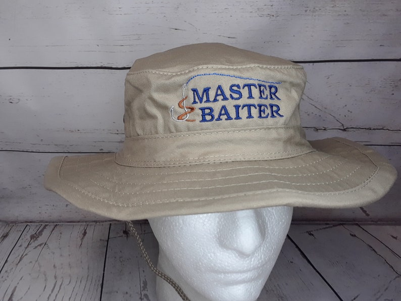 A khaki, wide-brimmed, bucket hat with a navy-blue text, a fish hook, and worm embroidered design that says 'MASTER BAITER'.