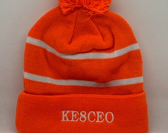 May include: Orange knit beanie with a white pom pom and white stripes. The beanie has the text "KESCEO" embroidered on the cuff.