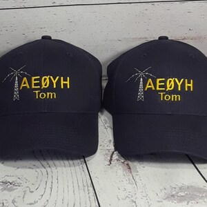 May include: Two dark navy blue baseball caps featuring identical embroidery. Each cap displays a white line-art graphic of a radio tower next to the yellow text AE&Oslash;YH and the name Tom embroidered in yellow lettering below the main text.