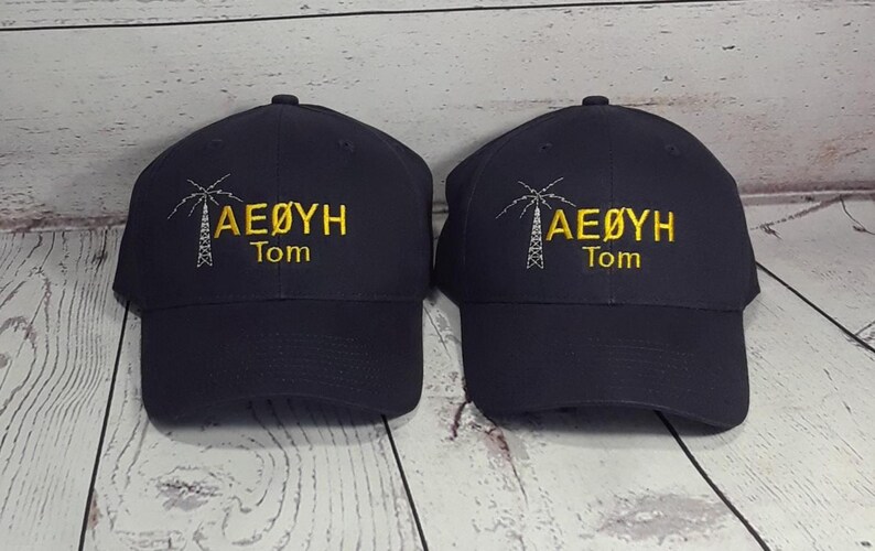 May include: Two dark navy blue baseball caps featuring identical embroidery. Each cap displays a white line-art graphic of a radio tower next to the yellow text AE&Oslash;YH and the name Tom embroidered in yellow lettering below the main text.