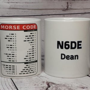 May include: Two white ceramic mugs with handles. The left mug features a red and black Morse Code chart with letters and numbers. The right mug displays the black text N6DE above the name Dean in a bold, sans-serif font on the front surface.