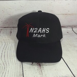 May include: A black baseball cap featuring an embroidered red radio tower icon on the left side. To the right of the icon, the text N2AHS is stitched in silver thread, with the name Mark embroidered in silver thread directly underneath it.
