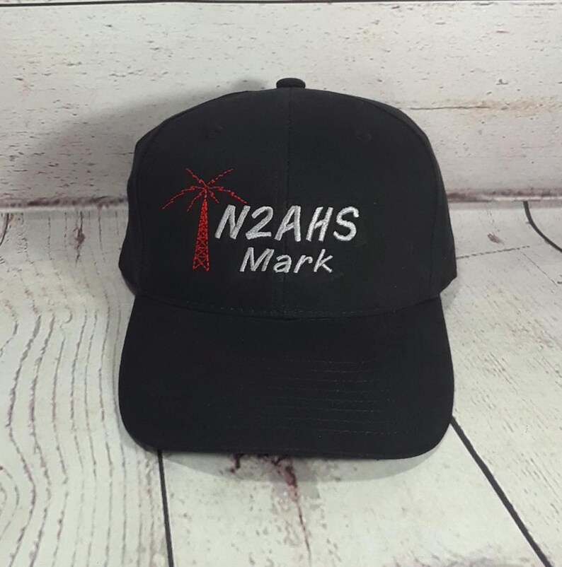 May include: A black baseball cap featuring an embroidered red radio tower icon on the left side. To the right of the icon, the text N2AHS is stitched in silver thread, with the name Mark embroidered in silver thread directly underneath it.