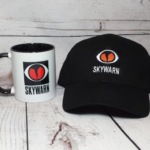 May include: A black and white mug with a red and orange eye logo and the text "SKYWARN" printed on it. Next to the mug is a black baseball cap with the same logo and text embroidered on it.