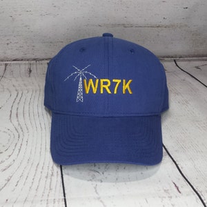 May include: A royal blue baseball cap featuring an embroidered white graphic of a radio tower and the yellow embroidered text WR7K. The cap has a structured crown, a curved brim, and a button on the top center, showcasing a clean design.