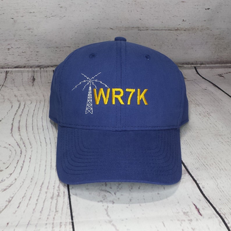May include: A royal blue baseball cap featuring an embroidered white graphic of a radio tower and the yellow embroidered text WR7K. The cap has a structured crown, a curved brim, and a button on the top center, showcasing a clean design.