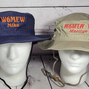 May include: Two sun hats with chin straps are displayed on mannequin heads. One hat is navy blue with "W6MEW Mike" embroidered in orange. The other is tan with "W6MEW-YL Marilyn" embroidered in red.