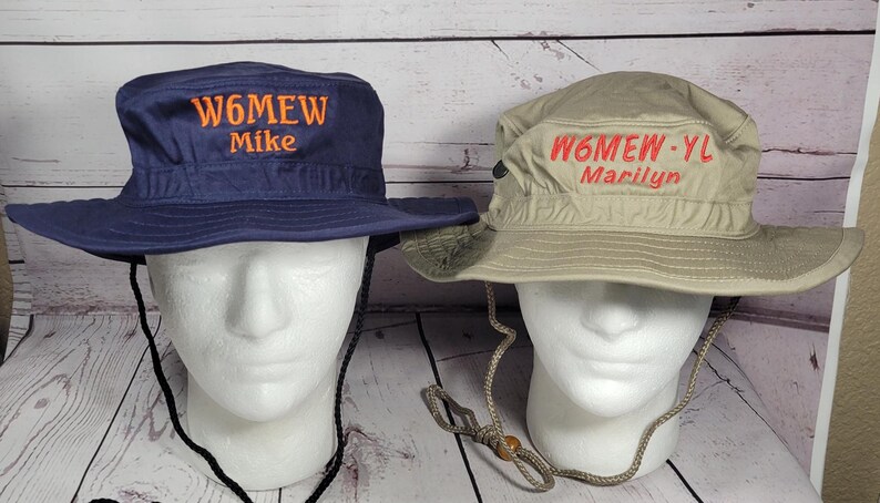 May include: Two sun hats with chin straps are displayed on mannequin heads. One hat is navy blue with "W6MEW Mike" embroidered in orange. The other is tan with "W6MEW-YL Marilyn" embroidered in red.