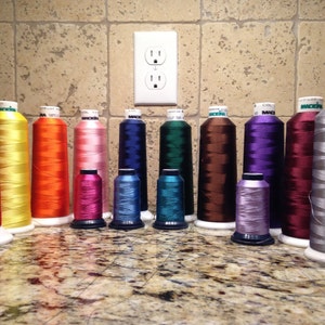 May include: A collection of Madeira embroidery thread spools in various colors including red, yellow, orange, pink, blue, green, brown, purple, maroon, and grey. The spools are arranged in two rows, with taller spools in the back and shorter spools in the front.