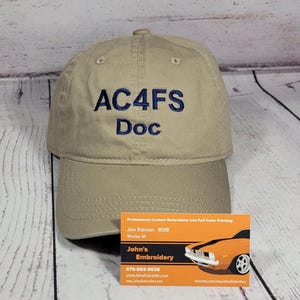 May include: A tan baseball cap featuring the embroidered text AC4FS Doc in dark blue thread. The cap has a structured crown, a curved brim with visible stitching, and eyelets for ventilation. The design is centered on the front panel of the hat.