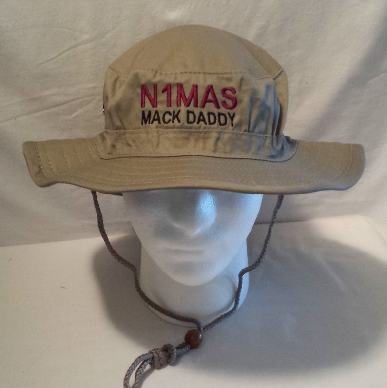 May include: A tan bucket hat with a wide brim and a chin strap. The hat features the embroidered text "N1MAS MACK DADDY" in pink thread across the front. The hat is displayed on a white mannequin head.