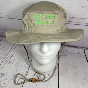 May include: A khaki-colored boonie hat with a wide brim and a chin strap. The hat features the embroidered text "KC1TRF JA5ELF" in green thread. The hat is displayed on a white foam head.