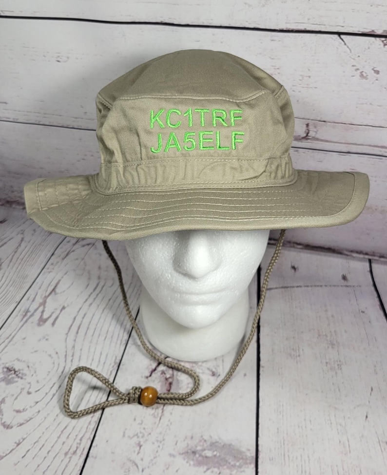 May include: A khaki-colored boonie hat with a wide brim and a chin strap. The hat features the embroidered text "KC1TRF JA5ELF" in green thread. The hat is displayed on a white foam head.