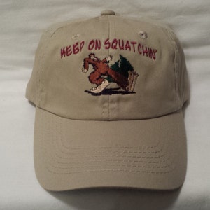 Op de afbeelding: Een beige baseballpet met een geborduurd beeld van een bruine Bigfoot die rent met een dennenboom. De tekst "KEEP ON SQUATCHIN'" is in rood boven het beeld geborduurd.