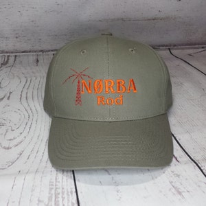 May include: An olive green baseball cap featuring an embroidered design in orange thread. The design includes a graphic of a transmission tower and the text N&Oslash;RBA Rod. The cap has a structured crown, a curved brim, and stitched eyelets on top.