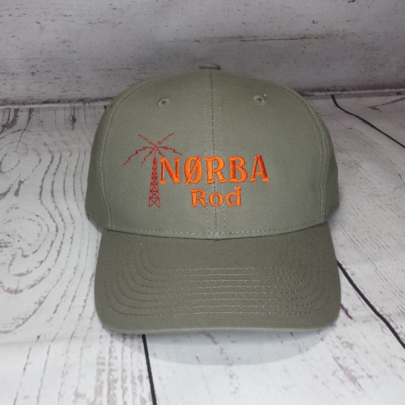 May include: An olive green baseball cap featuring an embroidered design in orange thread. The design includes a graphic of a transmission tower and the text N&Oslash;RBA Rod. The cap has a structured crown, a curved brim, and stitched eyelets on top.